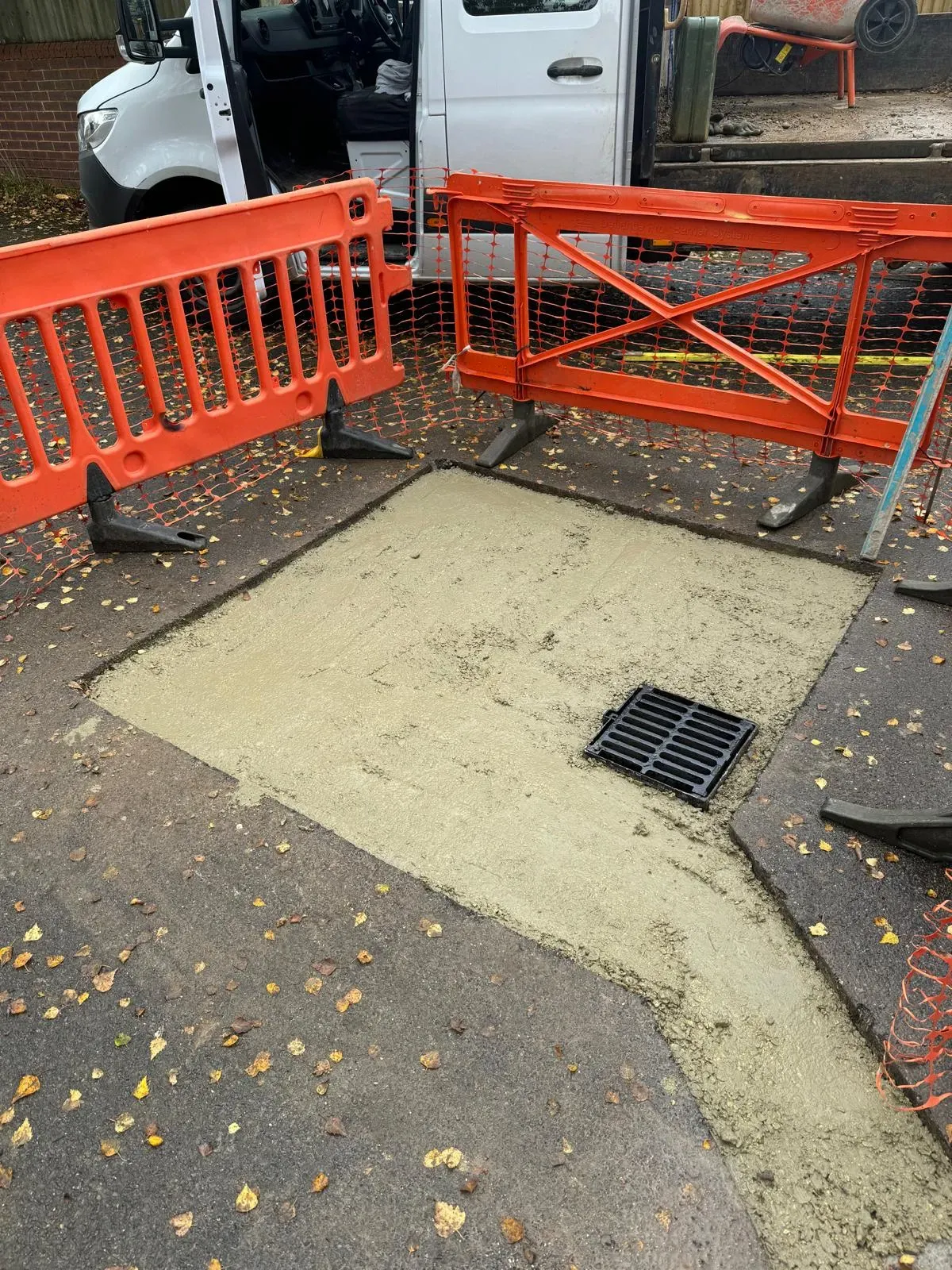 Newly installed storm drain grate in a fresh concrete patch on a paved surface, with orange safety barriers and a white work van in the background.
