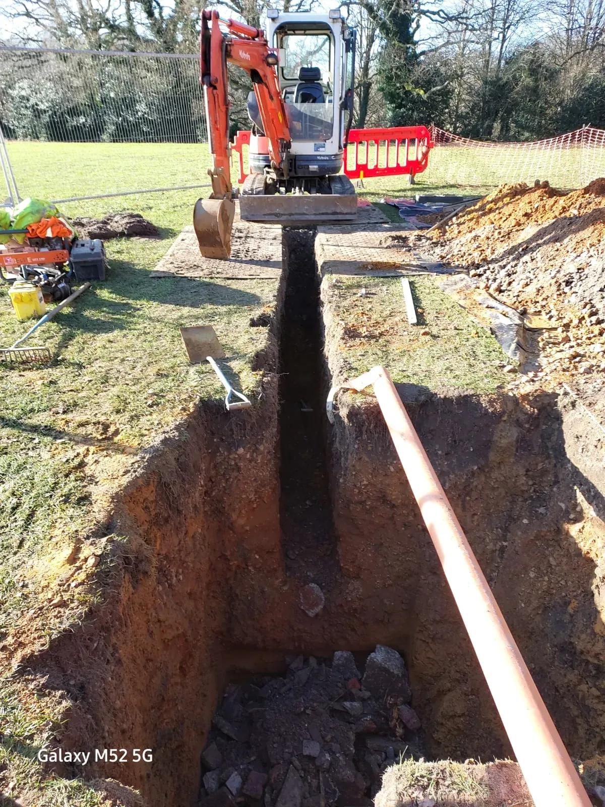 A mini excavator parked next to a deep trench with a light brown drainage pipe laid nearby, indicating active drain installation or repair work in a grassy field.