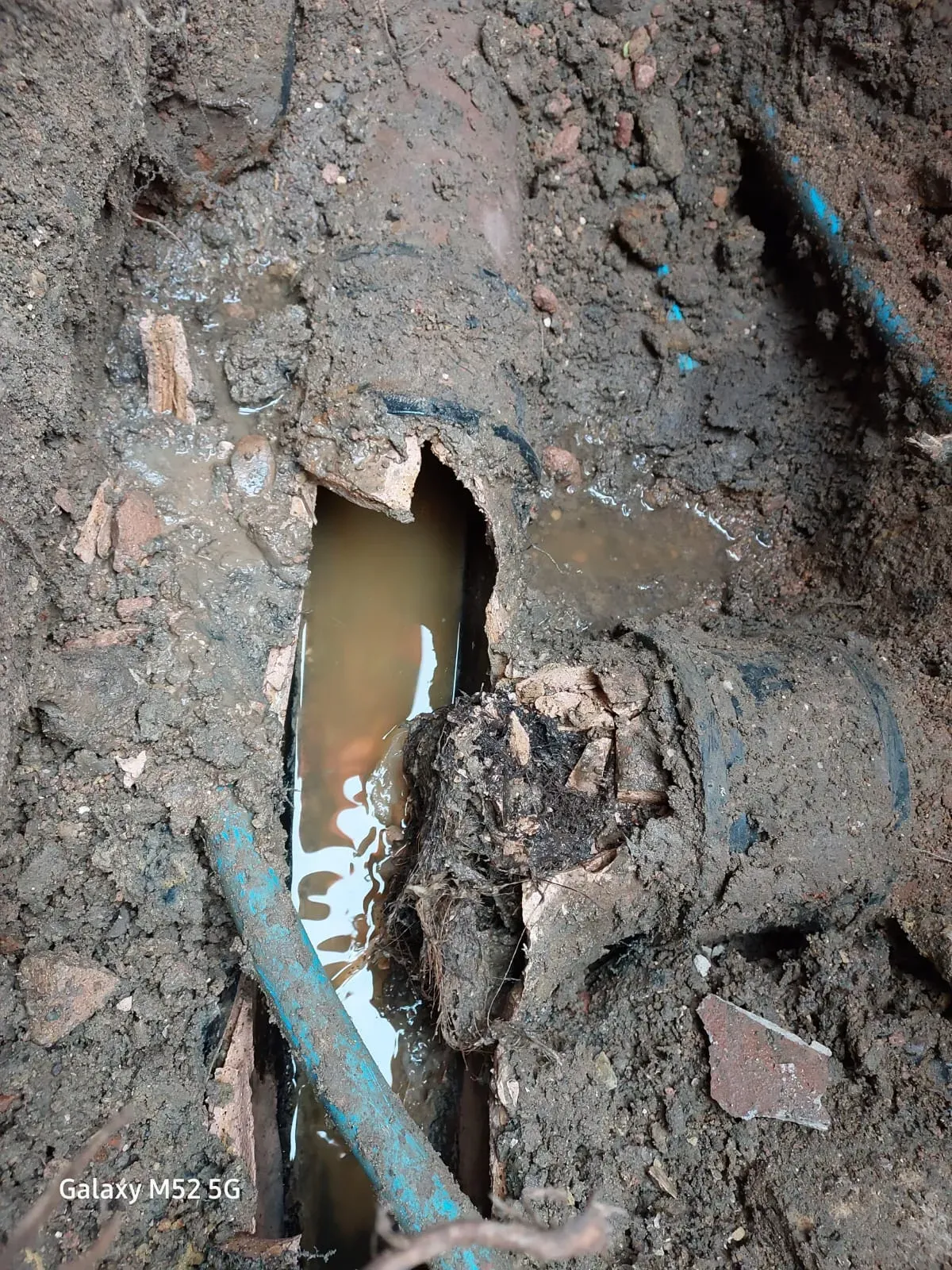 Severely damaged underground drain pipe with muddy water and debris in an excavated trench.